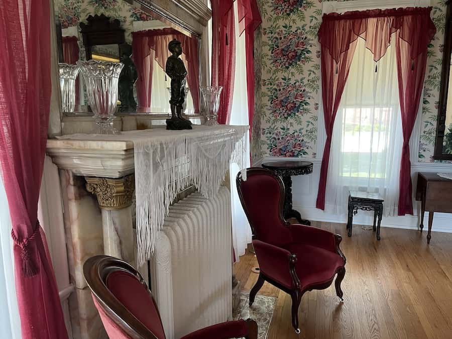 Inside the restored Schowengerdt House showing a mantle, fireplace, and red lace curtains near victorian red chairs.