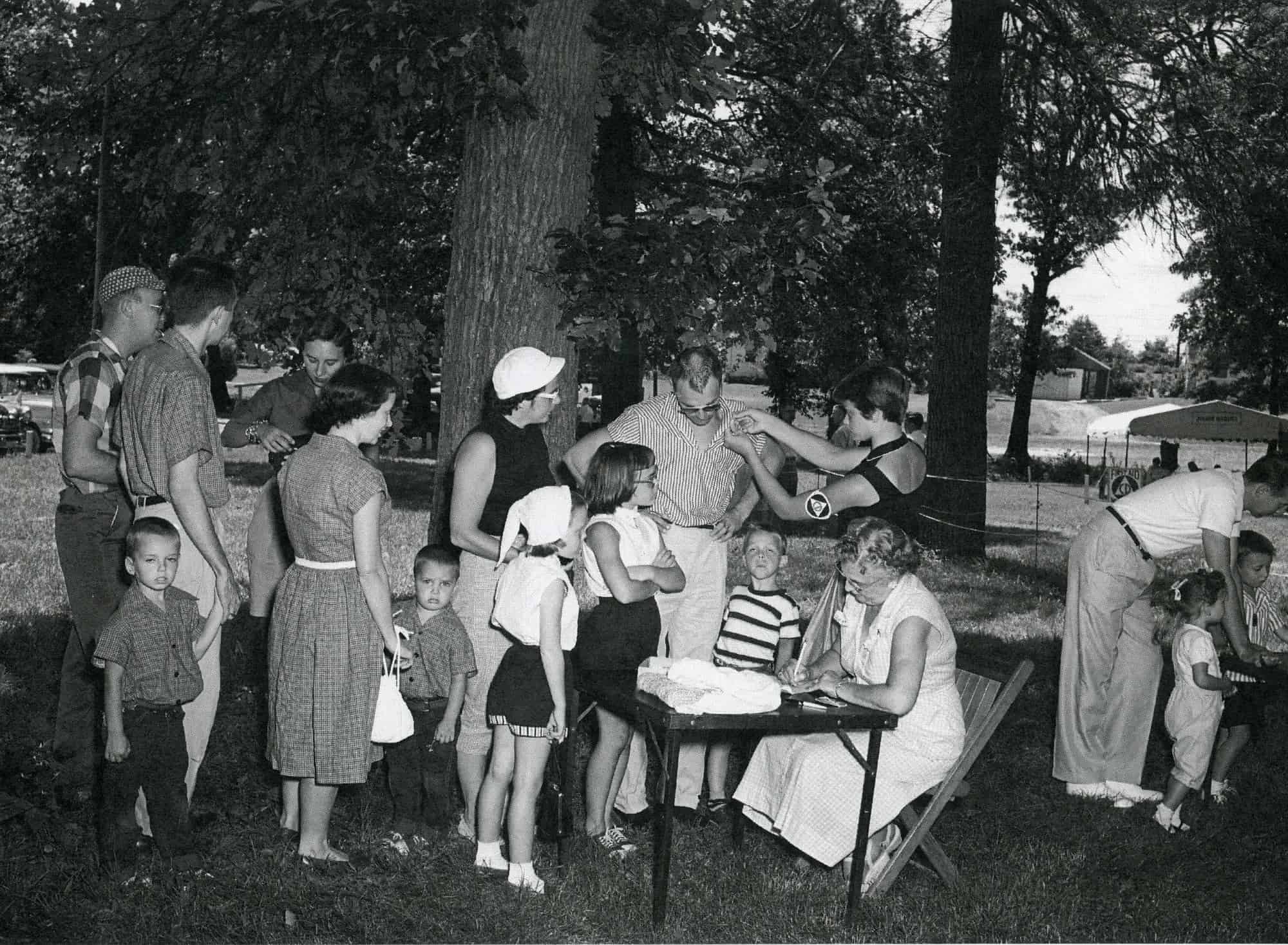 Parents and children line up at an outdoor table where a woman wearing a civil defense armband pins a tag on a man in a striped shirt while another woman writes at the table.