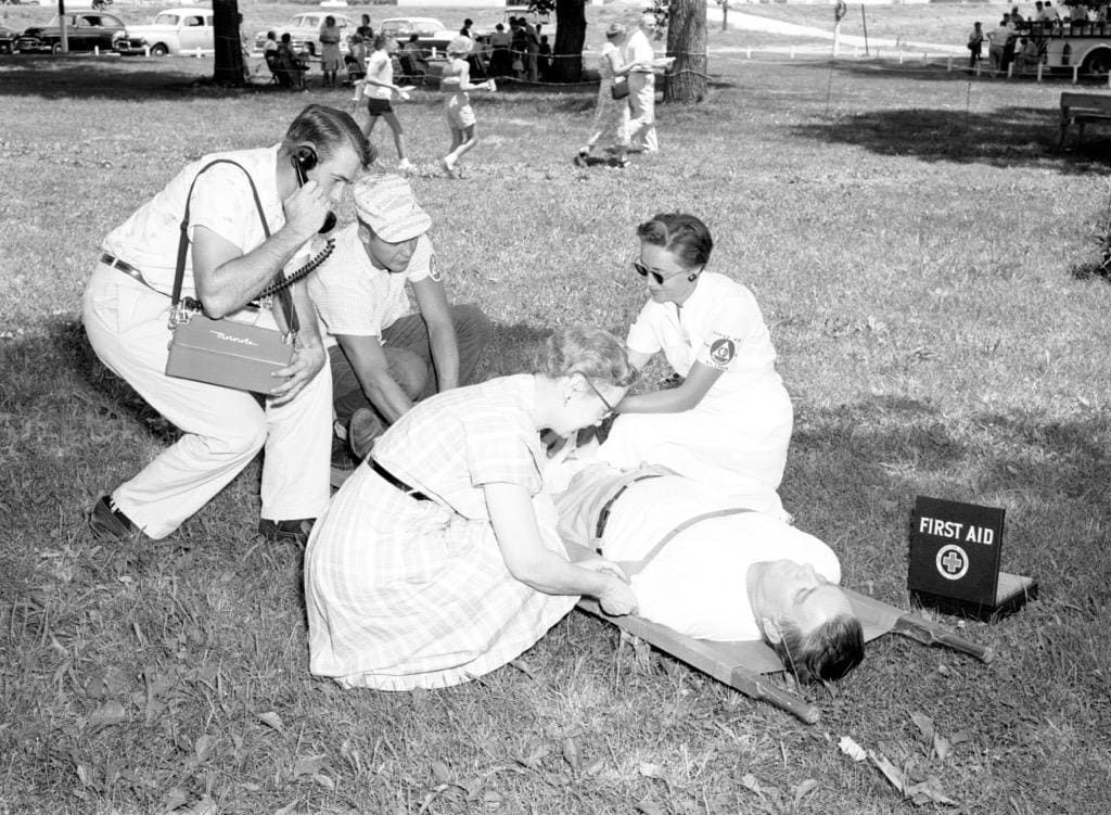 Volunteers wearing civil defense armbands kneel beside a man lying on a stretcher next to a First Aid box, while another volunteer talks on a portable radio.