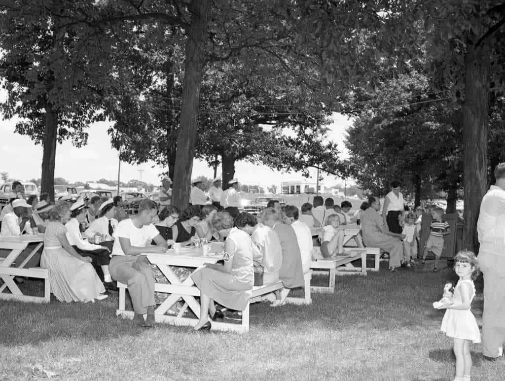 A crowd seated at white picnic tables under shade trees, including women in summer dresses, men in white uniform caps, and a small girl in a white dress standing in the foreground.