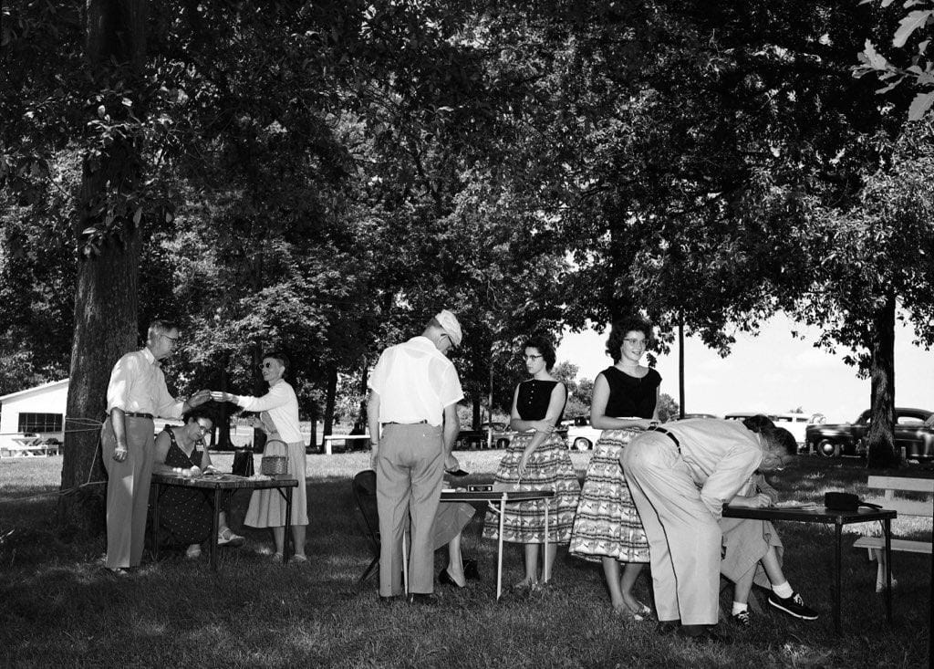 Volunteers seated at folding tables under large trees take down information from visitors, as two young women in dark tops and patterned skirts wait nearby.