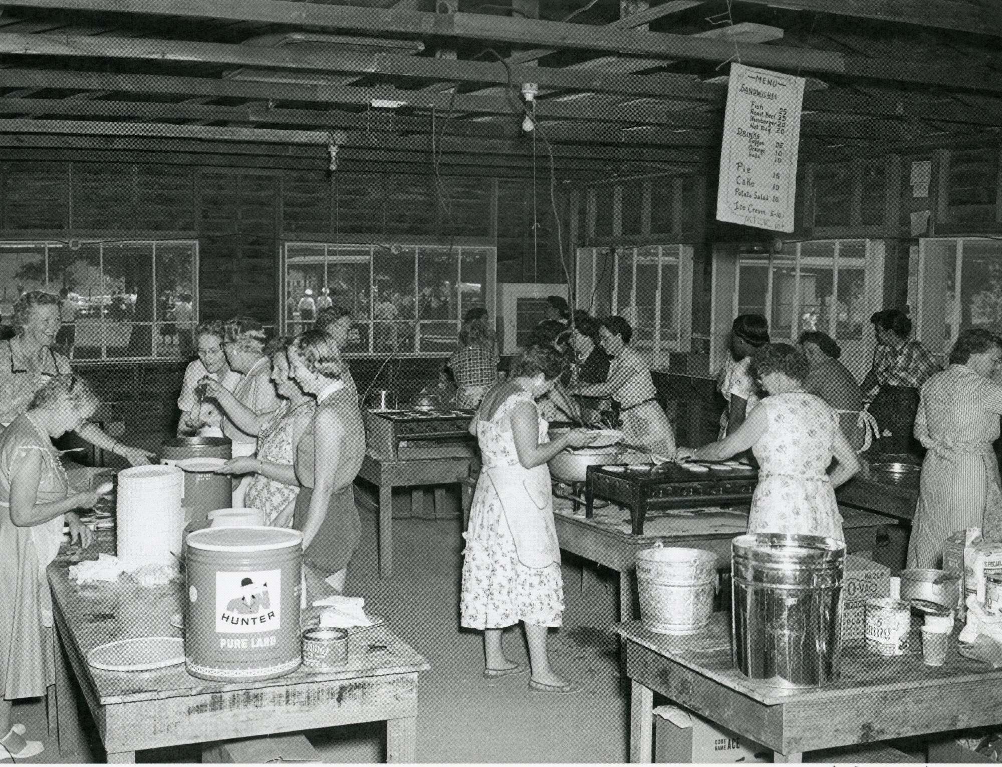 Women in aprons cook and serve food in a large wooden hall, with a can of Hunter pure lard on the table and a hand-lettered menu of sandwiches, drinks, pie, cake and ice cream hanging from the rafters.