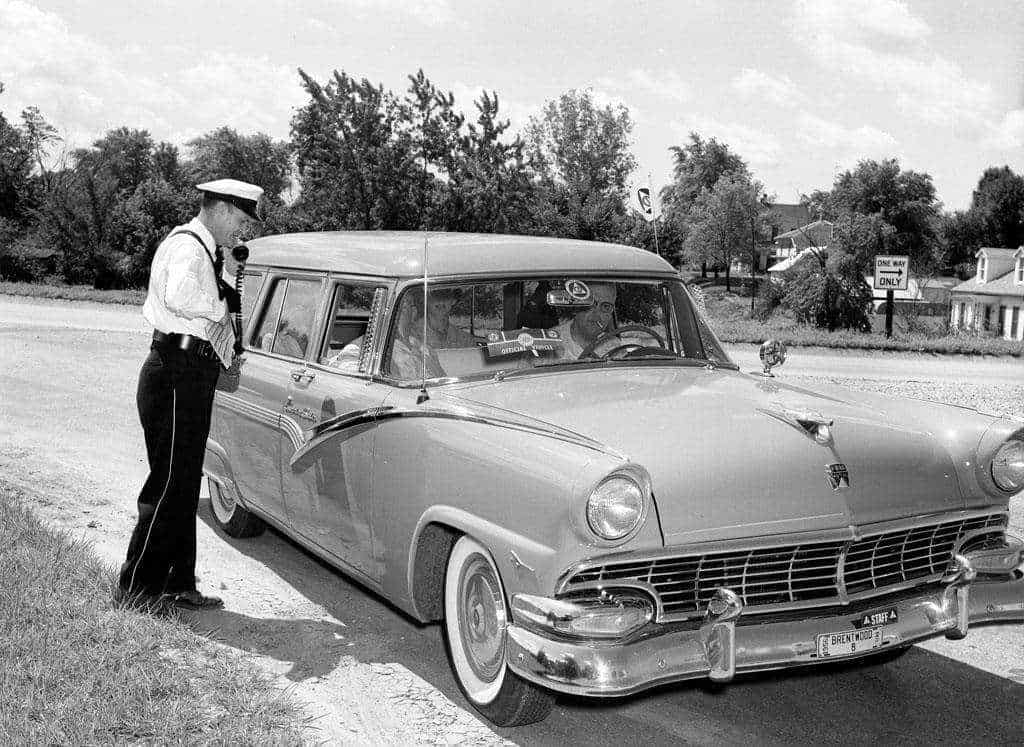 A uniformed officer in a white cap speaks into a radio handset beside a station wagon marked Official Vehicle with Brentwood staff license plates.