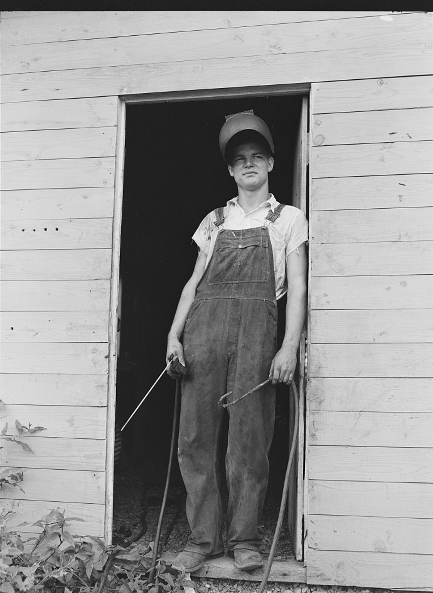 A farm boy in overalls and a cap stands in a barn doorway holding welding cables powered by Rural Electrification Administration electricity, Dunklin County, Missouri, 1942.