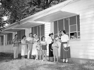 A group of women and children stand on the grass outside a white frame building with propped-open awning windows.