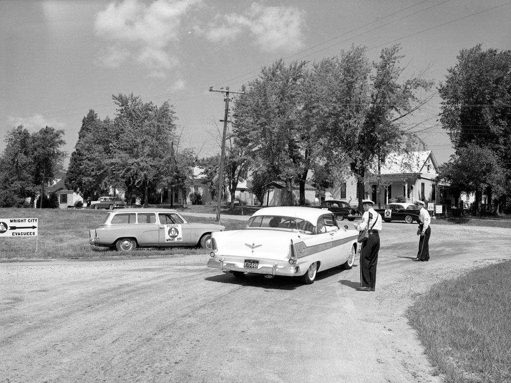 Two uniformed officers stop a two-tone 1950s sedan at a gravel intersection, near a Wright City police station wagon and a sign pointing to Wright City Evacuees.