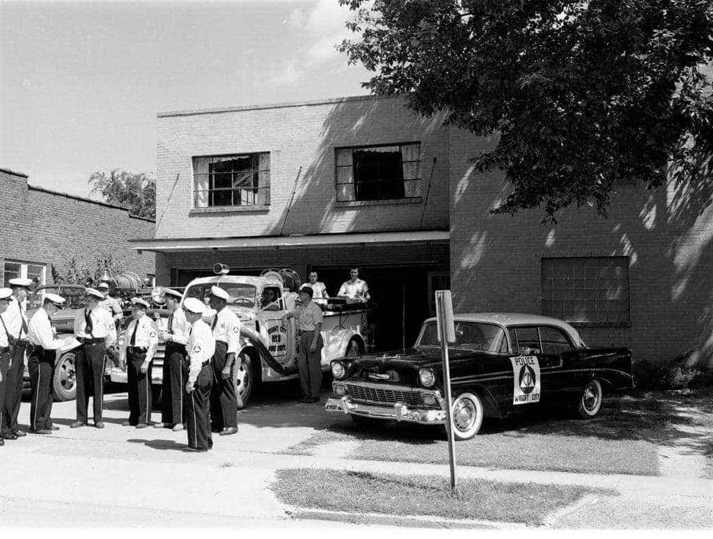 Uniformed officers gather outside a brick fire station beside a Wright City fire truck and a police car marked with the civil defense emblem.