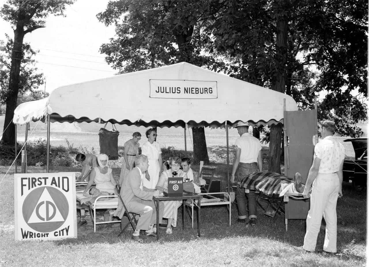 A first-aid station under a scalloped tent marked Julius Nieburg, with a sign reading First Aid, CD, Wright City. Nurses, a radio operator and bandaged patients on cots are inside.