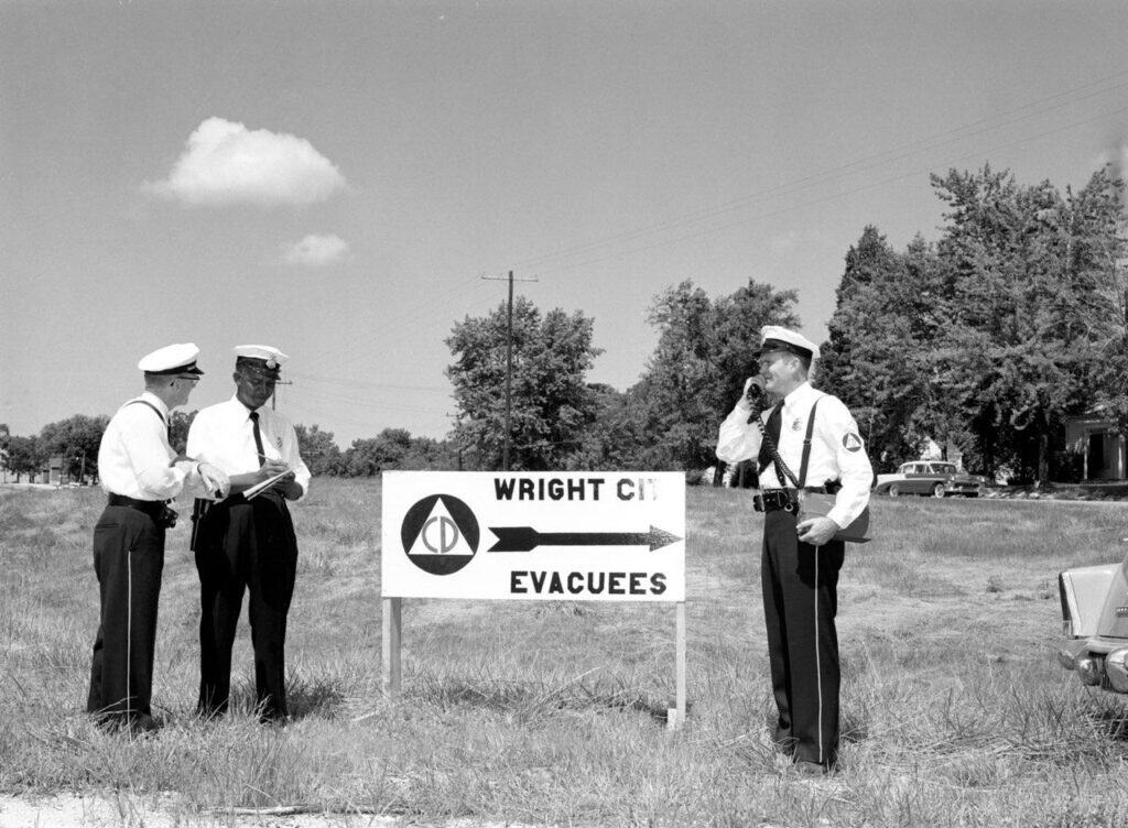 Three uniformed civil defense officers in white caps stand beside a sign with the CD emblem and an arrow reading Wright City Evacuees; one speaks into a radio handset.